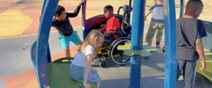 Kids enjoy We-Go-Round while sitting in a wheelchair, sitting on an interior bench, and running while pushing We-Go-Round from the outside.